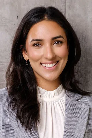 Portrait of a smiling woman with dark hair wearing a white blouse and gray blazer against a gray wall with a textured background.
