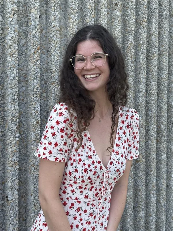 Julie Darcis Smiling woman with curly dark hair and round glasses, wearing a white dress with red flowers, standing before a textured gray wall.