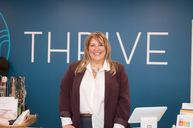 Susan Ingram Smiling woman in a white blouse and burgundy blazer stands at a reception desk in front of a blue wall with the word THRIVE behind her.