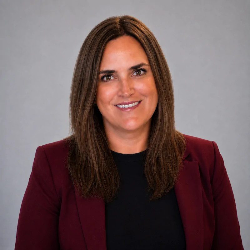 pic-Camila-Suvaric-new Professional headshot of a woman with straight brown hair, wearing a burgundy blazer and black top, smiling against a neutral gray background.